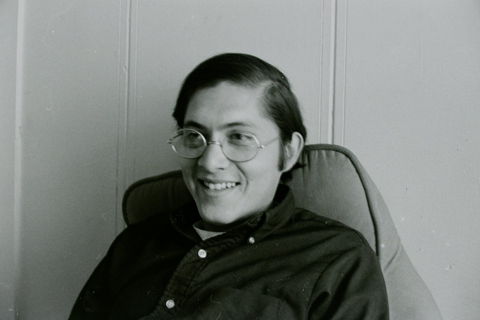 A young man in round wire glasses smiling in a chair